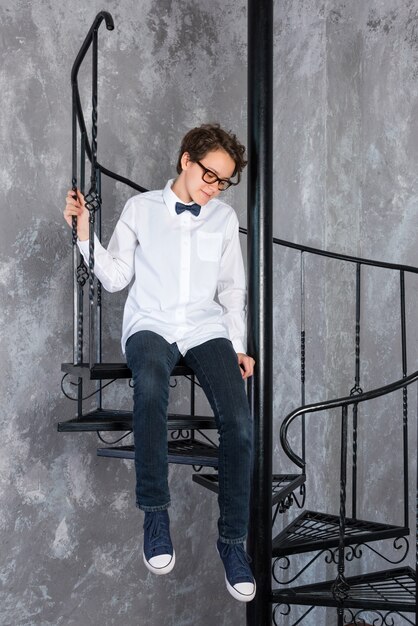 Adolescent intelligent portant des lunettes assis sur un escalier en colimaçon dans un loft appartement adolescente en blanc s...