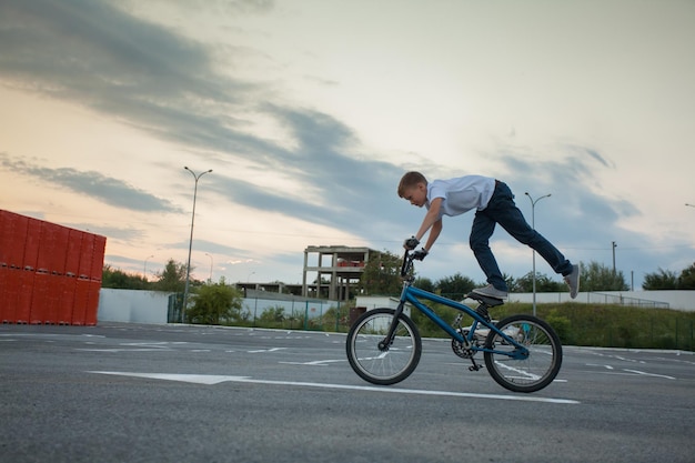 Adolescent intrépide faisant du vélo debout sur la selle sur une jambe. Tours spectaculaires du jeune motard sur le parking vide.