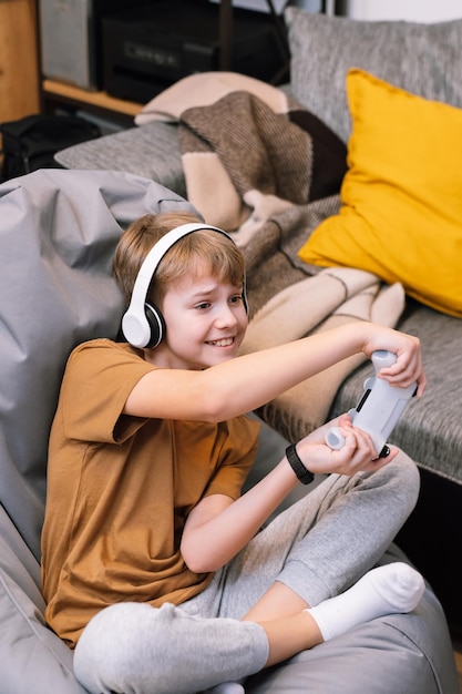 Adolescent jouant activement et imprudemment à un jeu vidéo avec un joystick assis sur un pouf sans cadre à la maison