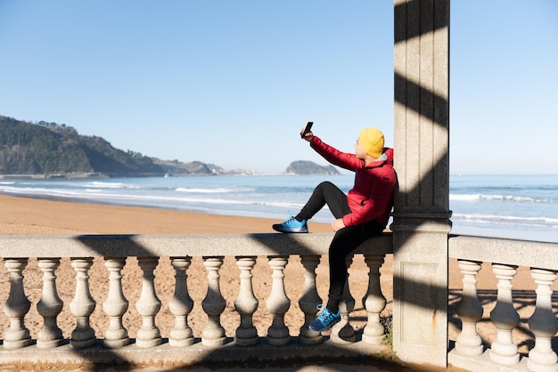 Adolescent latin prenant un selfie assis au bord de la mer.