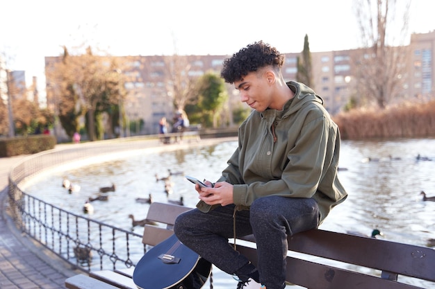 Adolescent latino assis sur un banc de parc avec un smartphone à la main