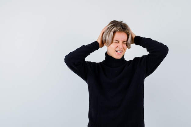 Adolescent mignon en pull à col roulé noir serrant la tête avec les mains et l'air irrité