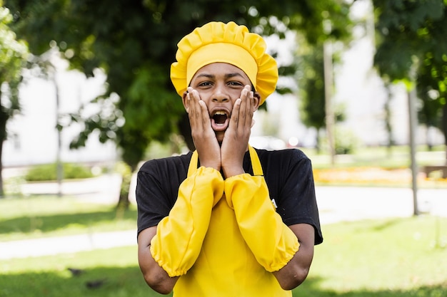 Adolescent noir africain choqué cuisinier en toque de chefs et uniforme de tablier jaune tenir les joues et surprendre Publicité créative pour café ou restaurant