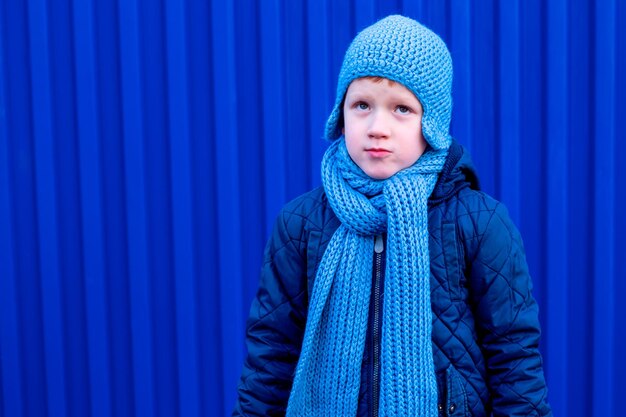 Adolescent rêvant réfléchi en chapeau bleu, écharpe, veste bleu foncé sur fond de mur bleu