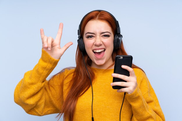Adolescent rousse femme sur mur bleu isolé écouter de la musique avec un mobile faisant un geste rock