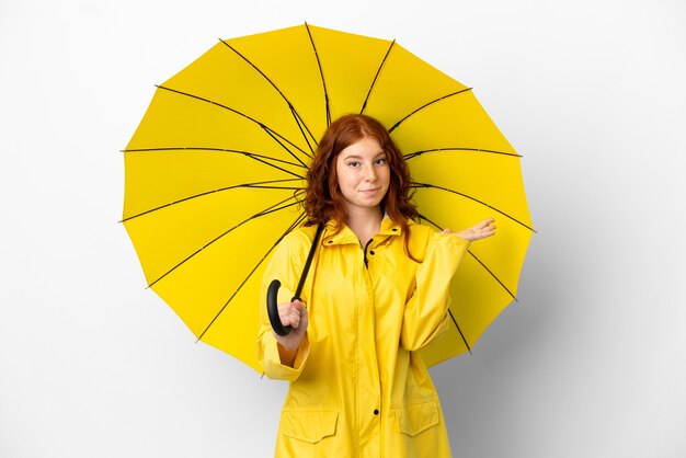 Adolescent rousse manteau imperméable et parapluie isolé sur fond blanc ayant des doutes tout en levant les mains