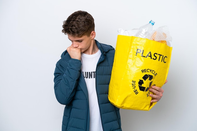 Adolescent russe tenant un sac plein de bouteilles en plastique à recycler sur fond blanc ayant des doutes