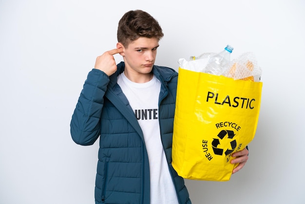 Adolescent russe tenant un sac plein de bouteilles en plastique à recycler sur fond blanc frustré et couvrant les oreilles