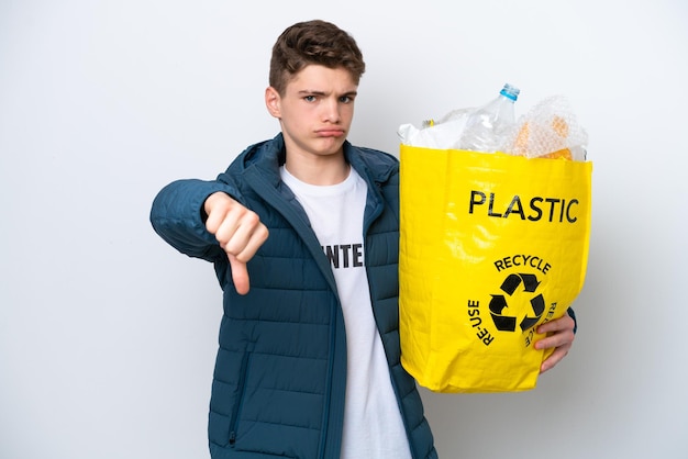 Adolescent russe tenant un sac plein de bouteilles en plastique à recycler sur fond blanc montrant le pouce vers le bas avec une expression négative