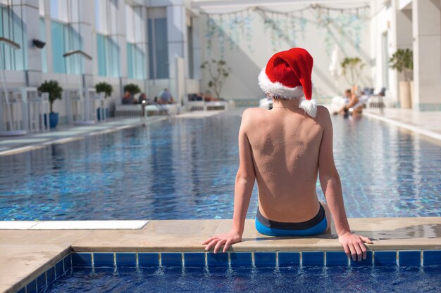 Adolescent sans visage en chapeau de père Noël rouge assis sur le bord de la piscine aux beaux jours