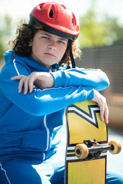 Photo adolescent sérieux dans un casque de protection s'appuyant sur la planche à roulettes à motifs et regardant devant lui