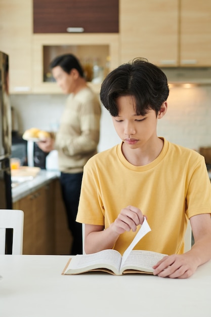 Adolescent sérieux, livre de lecture lorsque son père prépare le petit-déjeuner en arrière-plan