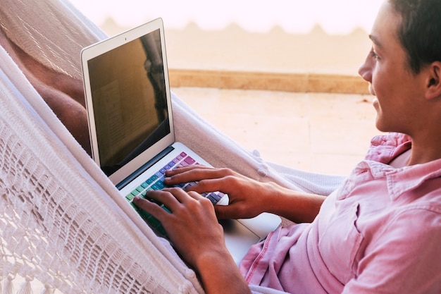 Adolescent seul en terrasse sur le hamac regardant des vidéos, travaillant ou jouant à des jeux vidéo avec son ordinateur portable ou son ordinateur et son téléphone - mode de vie en ligne et générations futures