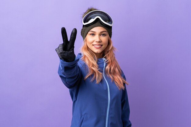 Adolescent skieur fille avec des lunettes de snowboard sur mur violet isolé souriant et montrant le signe de la victoire