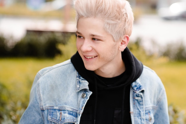 Un adolescent souriant de 16 à 17 ans aux cheveux blonds courts porte une veste en jean et un sweat à capuche noir