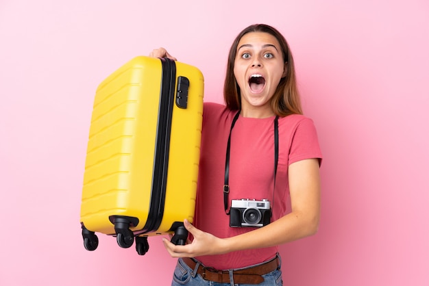 Adolescent voyageur fille tenant une valise sur rose isolé avec surprise et expression faciale choquée