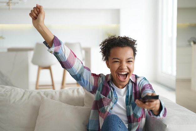 Une adolescente afro-américaine ravie célèbre son objectif en regardant la télévision assise sur un canapé à la maison