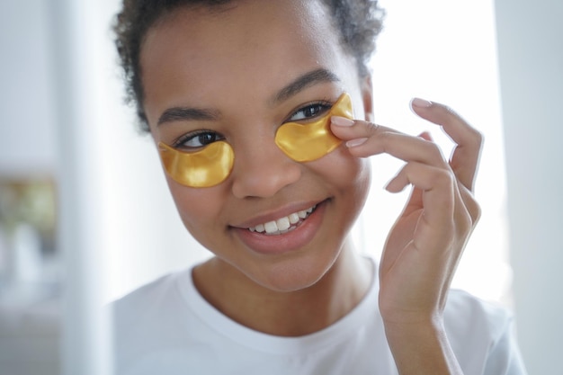 Une adolescente afro heureuse applique des pansements oculaires Hydratant pour la peau avec du sérum de collagène