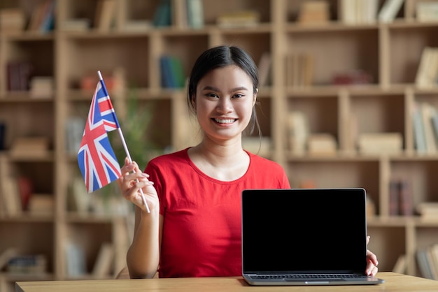 Une adolescente asiatique souriante tient le drapeau de l'angleterre près d'un ordinateur portable avec un écran vide à l'intérieur de la chambre