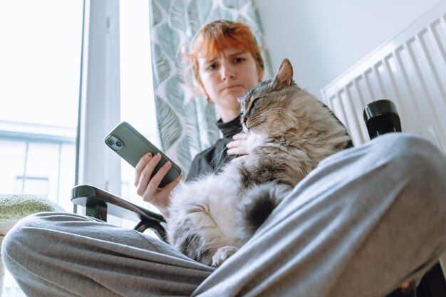 Une adolescente assise sur une chaise tenant un chat et regardant son téléphone.