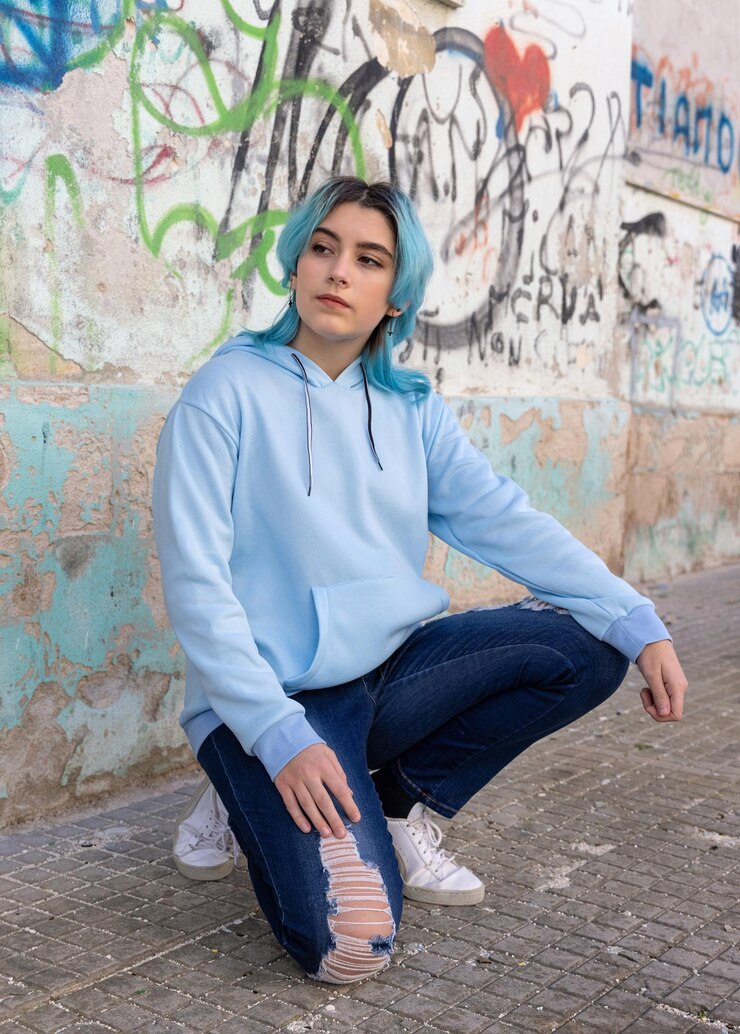 Photo adolescente aux cheveux bleus en sweat à capuche surdimensionné bleu clair et regardant de côté adolescente aux cheveux bleus assise sur ses hanches près du mur de graffitis maquette de vêtements