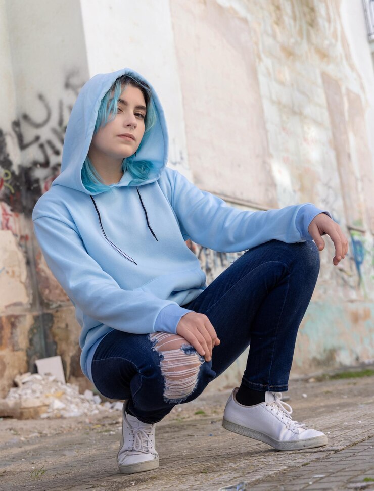 Photo adolescente aux cheveux bleus en sweat à capuche surdimensionné bleu clair et regardant de côté adolescente aux cheveux bleus assise sur ses hanches près du mur de graffitis maquette de vêtements
