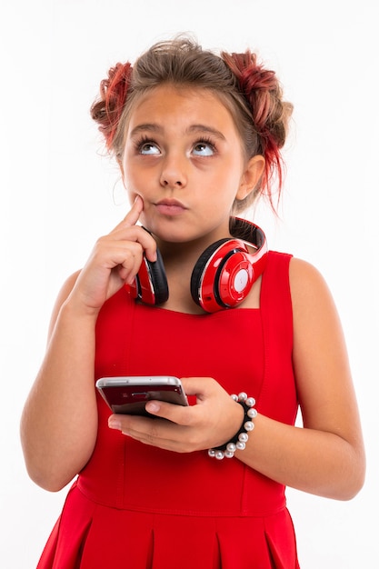 Adolescente aux longs cheveux blonds, pointes teintes en rose, farcies en deux touffes, en robe rouge, avec un casque rouge, un bracelet, debout et tenant le téléphone à la main et pensant