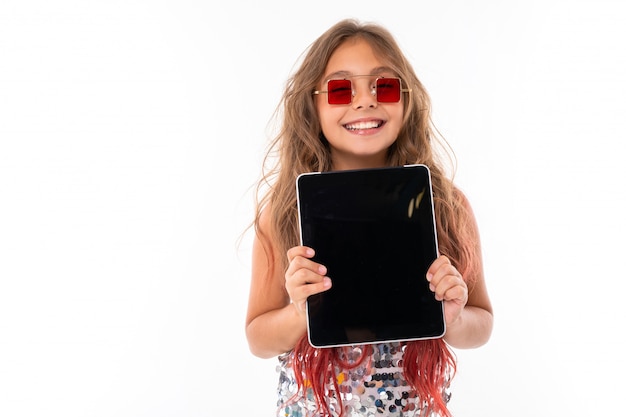 Adolescente aux longs cheveux blonds teints avec des pointes roses, en robe légère brillante, baskets en noir et blanc, lunettes, debout avec un casque, tenant une tablette dans ses mains