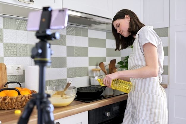 Adolescente, blogueuse culinaire cuisinant des crêpes à l'orange à la maison dans la cuisine, filmant une recette vidéo. Femelle avec bouteille d'huile près de la cuisinière. Loisirs, chaîne vidéo avec suiveurs, enfants et adolescents