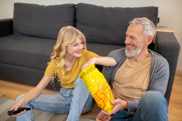 Une adolescente blonde assise par terre avec son père et mangeant des chips