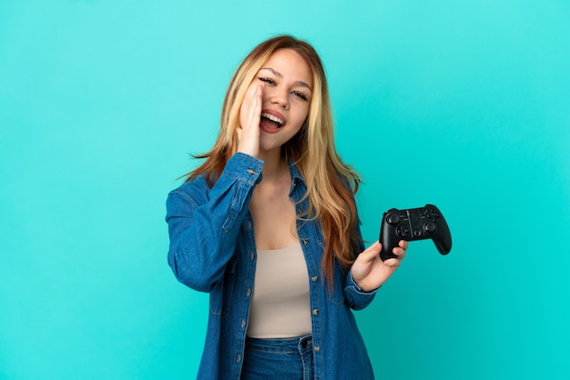 Adolescente blonde jouant avec un contrôleur de jeu vidéo sur un mur isolé criant avec la bouche grande ouverte