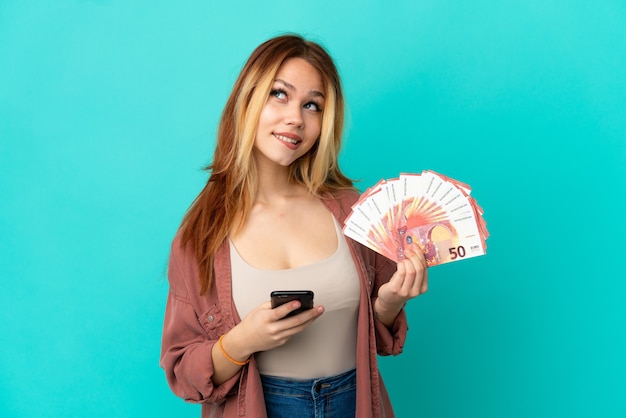 Adolescente blonde prenant beaucoup d'euros sur un mur bleu isolé tenant du café à emporter et un mobile tout en pensant à quelque chose