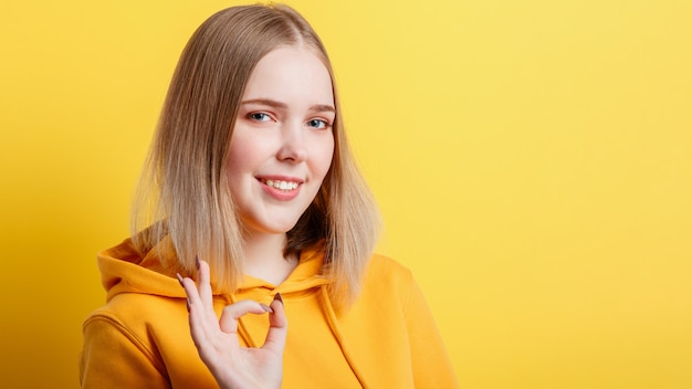 Une Adolescente Blonde Souriante Et Calme Montre Un Symbole De Geste Ok Positif Comme Et Une Approbation Isolée Sur Fond Jaune De Couleur. Portrait Jeune Femme Montre Un Geste De La Main Ok. Longue Bannière Web Avec Espace De Copie.