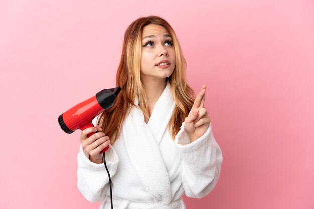 Adolescente blonde tenant un sèche-cheveux sur fond rose isolé avec les doigts croisés et souhaitant le meilleur