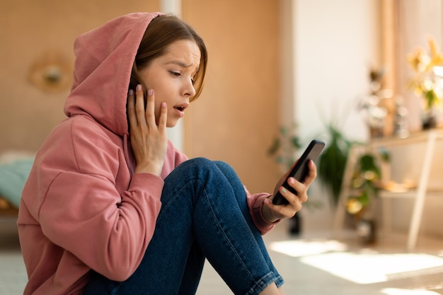 Une Adolescente Bouleversée Et Choquée Lisant Un Message De Mauvaise Nouvelle Sur Un Téléphone Portable Touchant La Joue Avec étonnement Assis à La Maison