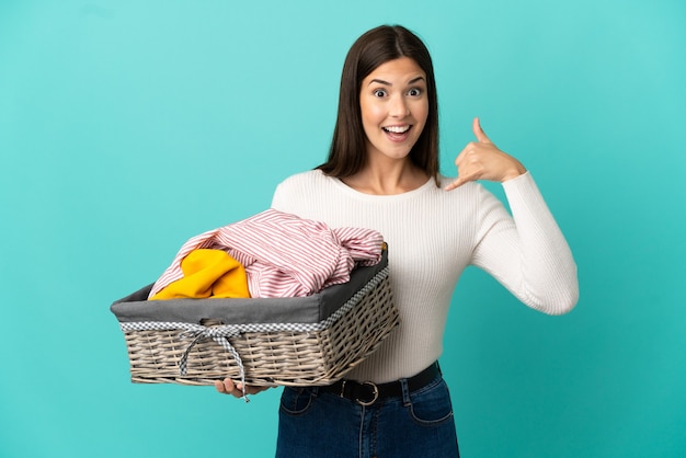 Adolescente brésilienne tenant un panier de vêtements isolé sur fond bleu faisant un geste de téléphone. Rappelle-moi signe