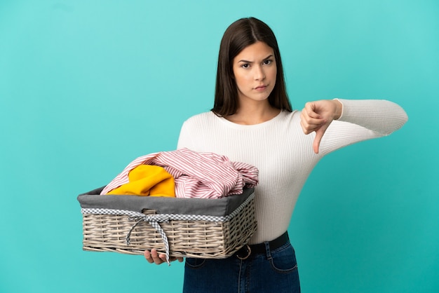 Adolescente brésilienne tenant un panier de vêtements isolé sur fond bleu montrant le pouce vers le bas avec une expression négative