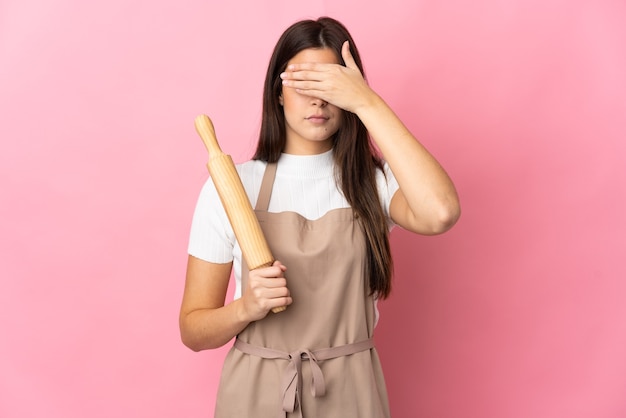 Adolescente brésilienne tenant un rouleau à pâtisserie isolé sur fond rose couvrant les yeux par les mains. Je ne veux pas voir quelque chose