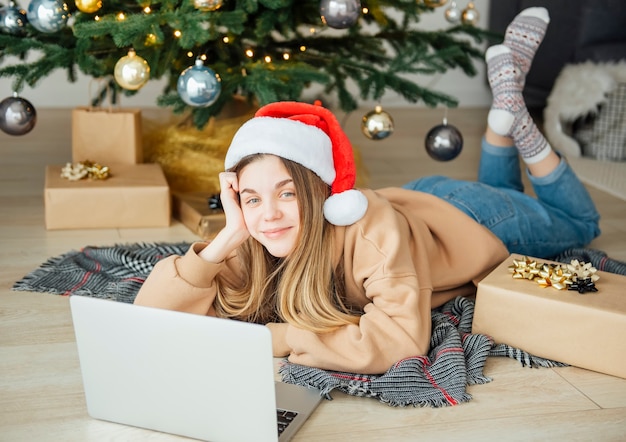 Adolescente avec des cadeaux et un ordinateur portable près de l'arbre de Noël. Intérieur du salon avec sapin de Noël et décorations. Nouvelle année. Donner en cadeau.