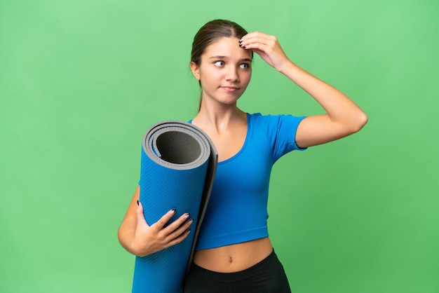 Adolescente caucasienne allant à des cours de yoga tout en tenant un tapis sur fond isolé ayant des doutes et avec une expression de visage confuse