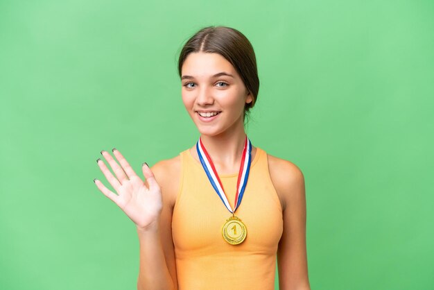 Adolescente caucasienne avec des médailles sur fond isolé saluant avec la main avec une expression heureuse