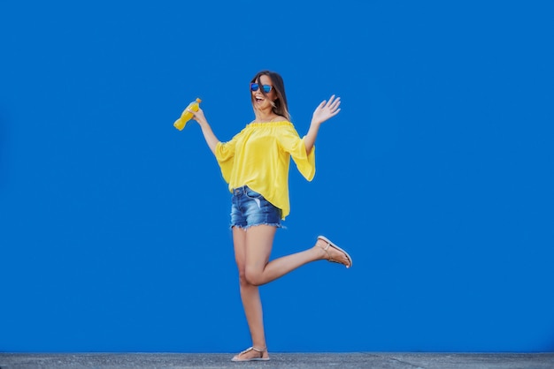 Adolescente caucasienne souriante en chemisier jaune et avec des lunettes de soleil tenant du jus d'orange en se tenant devant le mur bleu.