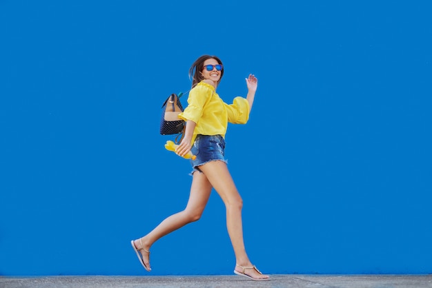 Adolescente caucasienne souriante en chemisier jaune et avec des lunettes de soleil tenant le jus d'orange en courant devant le mur bleu.