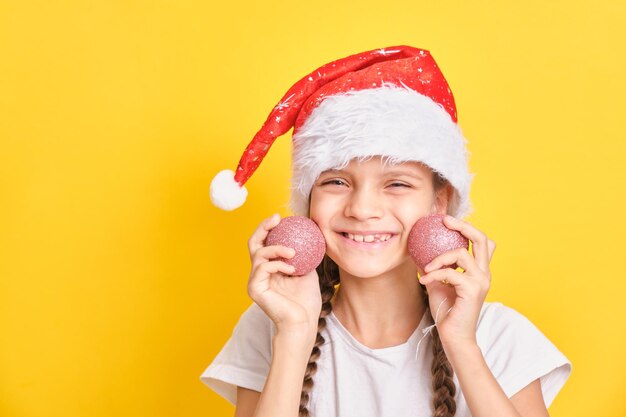 Adolescente en chapeau de père Noël tenant des boules de Noël sur fond jaune