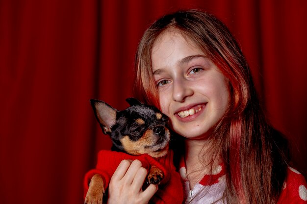 Adolescente et un chihuahua vêtu d'un pull rouge pour chiens à la maison sur fond rouge.