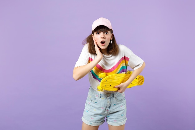 Une adolescente choquée dans des vêtements vifs tient une planche à roulettes jaune en gardant la bouche ouverte en mettant la main sur la joue isolée sur fond pastel violet. Émotions sincères des gens, concept de style de vie. Maquette de l'espace de copie.