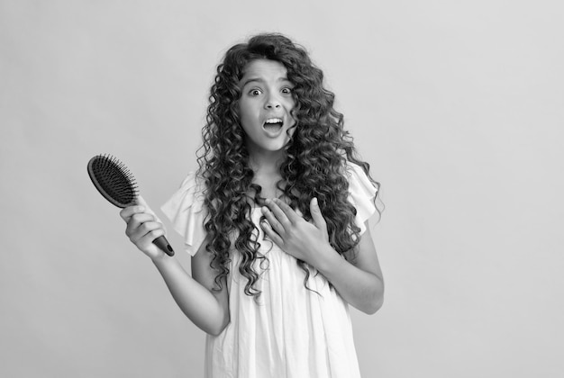 Adolescente choquée avec de longs cheveux bouclés tenant une brosse à cheveux en peigne pour peigner les habitudes quotidiennes