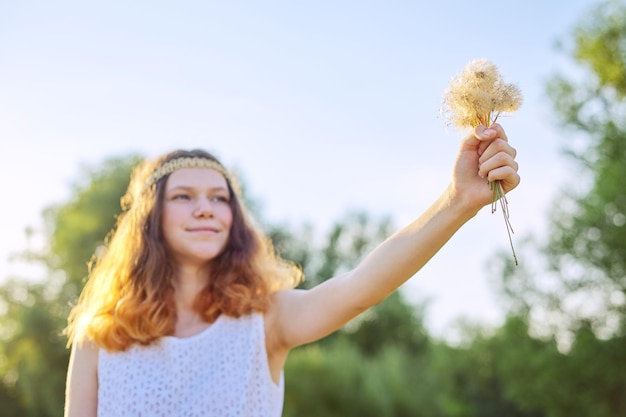 Adolescente avec une coiffure hippie tenant des pissenlits secs dans sa main. Vacances d'été, vacances, nature, beauté, style de vie. Ciel, prairie, fond d'éclairage au coucher du soleil