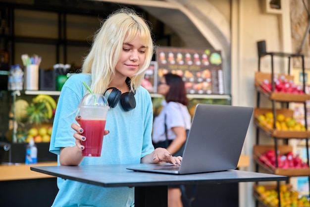 Adolescente dans un bar à jus de fruits en plein air de fruits buvant du jus de vitamine frais à l'aide d'un ordinateur portable