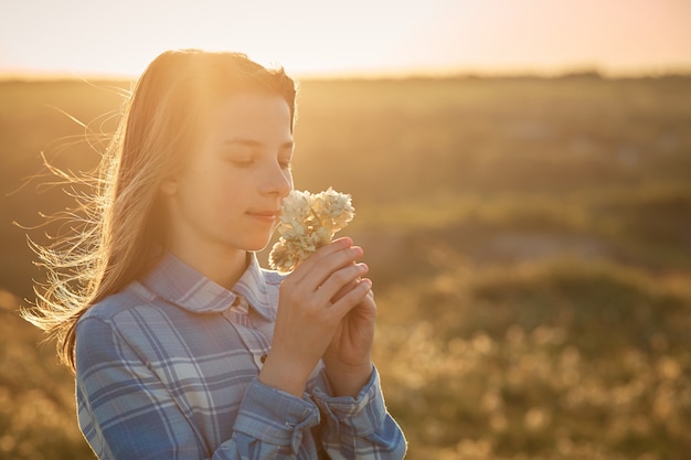 Une adolescente dans un champ au coucher du soleil avec des fleurs dans ses mains.